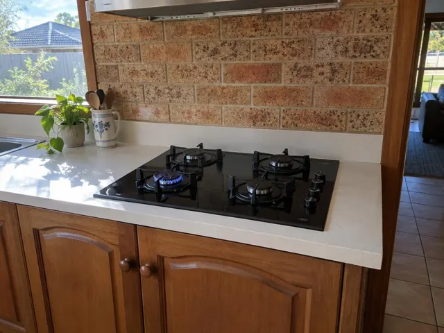 Black gas cooktop set into a white benchtop with brick backsplash and timber cabinets in Fern Bay