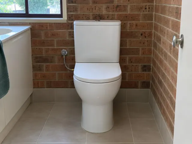 White toilet in a tiled bathroom with brick walls and a vanity beside a window in Fullerton Cove